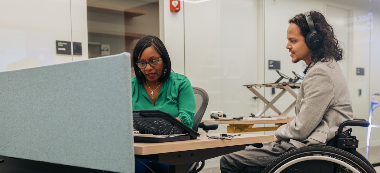 Photo by Jordan Nicholson, CC-BY-ND 4.0 Two people sitting at a desk, one wearing headphones.