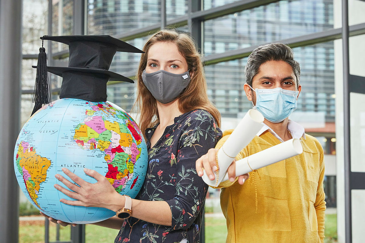 Zwei Personen mit Doktorhüten und gerolltem Papier in der Hand, welche die Abschlussarbeiteten darstellen sollen. Beide haben eine Atemschutzmaske auf und die Person links hält einen Globus in der Hand.